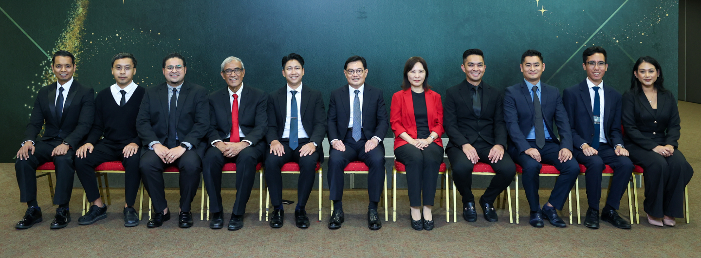 Eleven people in suits sit in a row against a dark green background.
